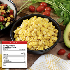A bowl of yellow freeze-dried corn kernels on a wooden table, with a partially visible salad bowl, cherry tomatoes, cilantro, and an avocado in the background. A product label overlay on the left reads 'Freeze-Dried Corn Nutrition Facts' and '1 Pouch Included'.